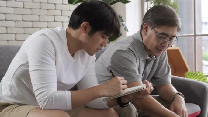 快乐的两代亚洲男子家庭老父亲写纸，而年轻的成年儿子使用数字笔记本电脑观看社交媒体坐在沙发上。资深父子