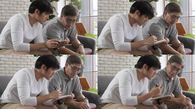 快乐的两代亚洲男子家庭老父亲写纸，而年轻的成年儿子使用数字笔记本电脑观看社交媒体坐在沙发上。资深父子