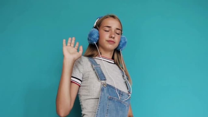 可爱的十几岁的女孩在室内蓝色背景的耳机里跳舞，随着节奏的变化听音乐。女孩微笑着，戴着耳机听音乐。一个