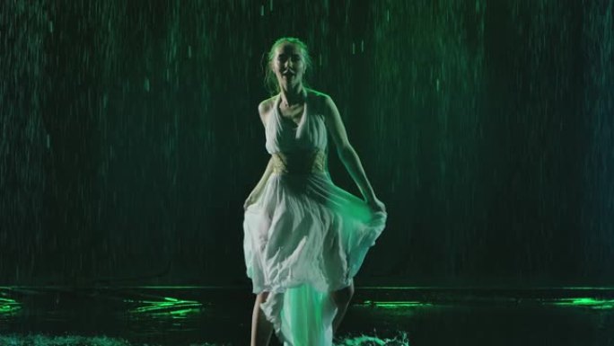 一位美丽的年轻女子穿着湿漉漉的裙子，在雨点下热情地跳舞。深色背景和工作室灯光。缓慢的运动。近距离