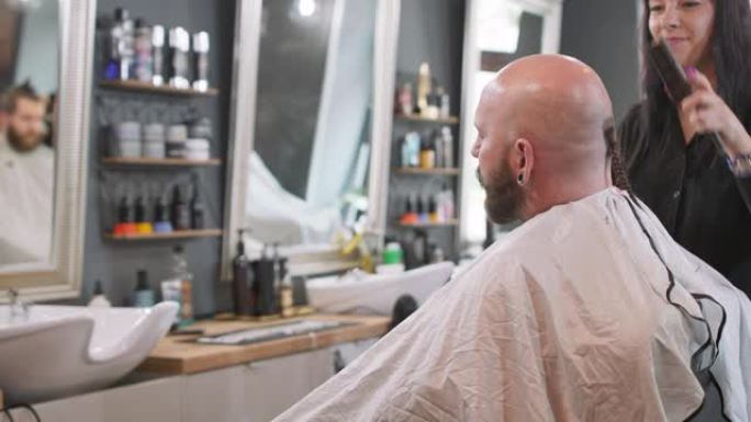 一位女理发师转向镜头，刷胡子男人光滑剃光的没有头发的头，他正坐在理发师的椅子上，面对镜子，说话。背景