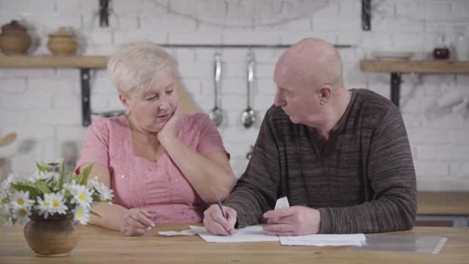 白人老年夫妇处理账单的肖像。秃头男人和金发女人坐在桌子旁聊天。高级配偶解决他们的收入和支出。财务，退