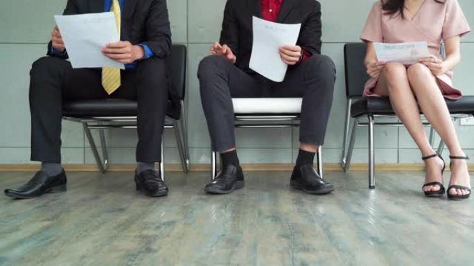 员工腿特写镜头，失业的商业人士申请人小组坐在排队排的椅子上等待他们的公司面试。他们看起来压力很大，焦