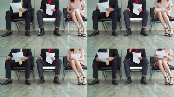 员工腿特写镜头，失业的商业人士申请人小组坐在排队排的椅子上等待他们的公司面试。他们看起来压力很大，焦