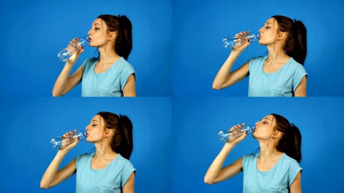 深色长发的黑发女孩在工作室蓝色背景下锻炼后，正在喝塑料透明瓶子里的纯净水