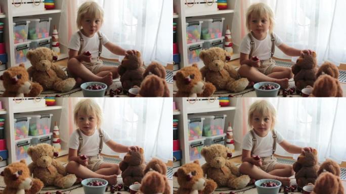 蹒跚学步的金发孩子，可爱的男孩，在家用泰迪熊吃樱桃