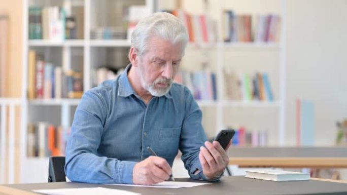 认真的老商人使用智能手机并在图书馆做文书工作