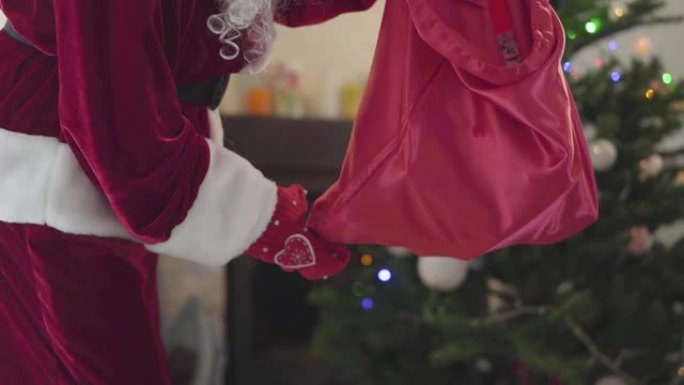 穿着圣诞老人服装的面目全非的男人在新年树前把礼物从一个袋子摇到一个地板。节日快乐、传统、圣诞节的概念