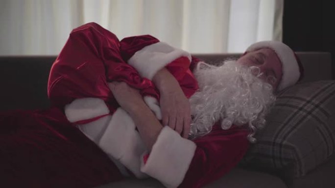 醉酒的老人，假白胡子，穿着圣诞老人的服装，睡在家里的沙发上，抱着礼物的包。坏圣诞老人。酗酒，消极主义
