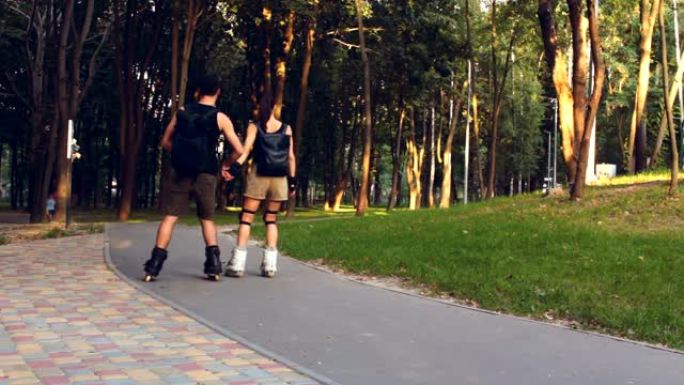 年轻幸福的夫妇牵着手在公园里溜旱冰。年轻迷人的女孩和一个年轻的家伙在公园里溜冰。恋爱中的年轻夫妇在夏