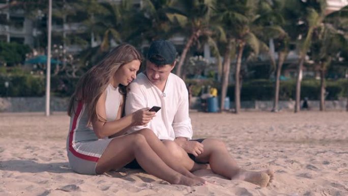赤脚夫妇坐在沙滩上，女孩坐在智能手机上