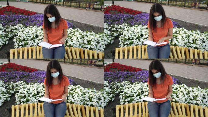 一个戴着医用口罩的女孩在长凳上看书。社交远程，远程教育。一个学生正在公园里准备上课。一个留着黑色长发