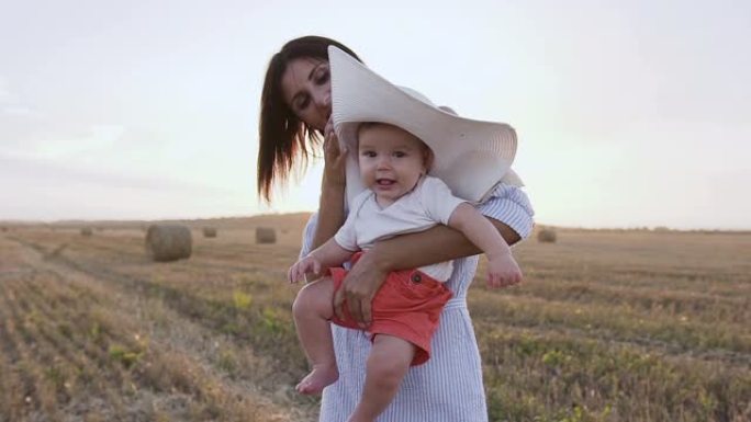 一位戴着大帽子的婴儿在夕阳下与母亲一起走在田野里的肖像。快乐的年轻女子带着小儿子在夏天的晚上在野外玩