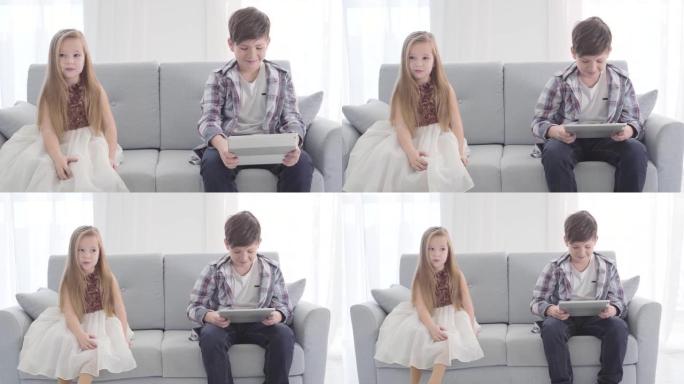 高加索小男孩看着平板电脑屏幕的肖像，坐在他旁边的可爱的女孩害羞地瞥了一眼她的爱。童年，初恋。