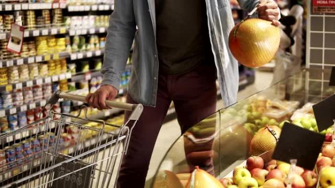 在一家杂货店超市的农产品部门，穿着蓝色衬衫的无法辨认的男人在购买水果和蔬菜的镜头。带着柚子，把它放进