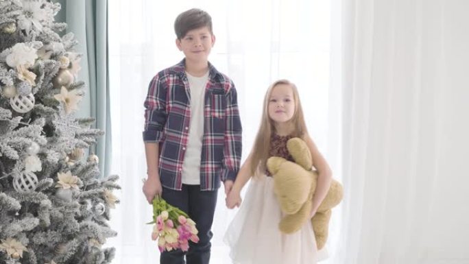 白人男孩和女孩的肖像站在圣诞树旁边，握手。黑发男孩握着初恋的手和一束郁金香，小女士拿着泰迪熊。