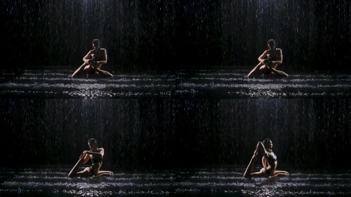 美丽的黑发年轻女子在雨中表演复杂的体式。工作室的光线反射在水面上发光。在黑色背景的黑暗工作室拍摄。慢
