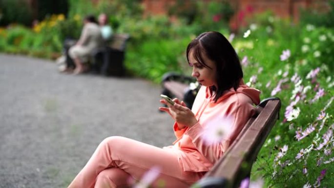年轻女子坐在公园的长凳上，用智能手机翻转新闻。成年女性在新鲜空气中享受手机休息