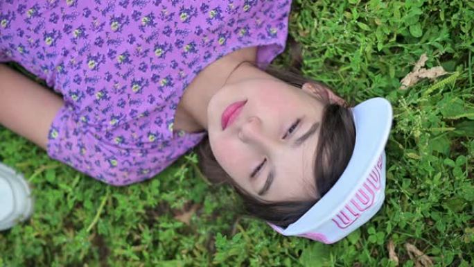 一个戴着帽子和粉红色t恤的可爱的少女躺在草地上，微笑着