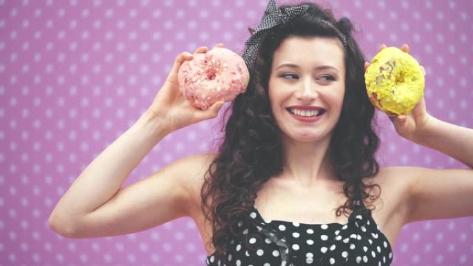 可爱的年轻卷发女孩站着，举起两个粉红色和黄色糖霜的美味甜甜圈，达到耳朵的水平。