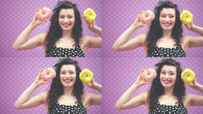 可爱的年轻卷发女孩站着，举起两个粉红色和黄色糖霜的美味甜甜圈，达到耳朵的水平。