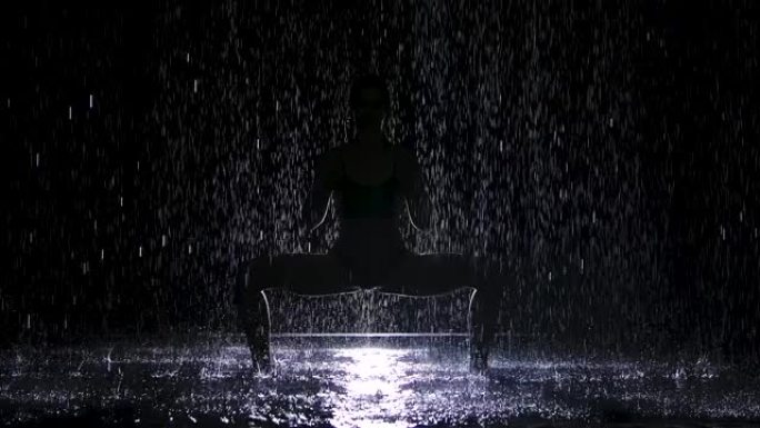 在昏暗的工作室里冒着倾盆大雨做瑜伽练习。一个白色的聚光灯照亮了水面和一个年轻女子的身体。轮廓。慢动作