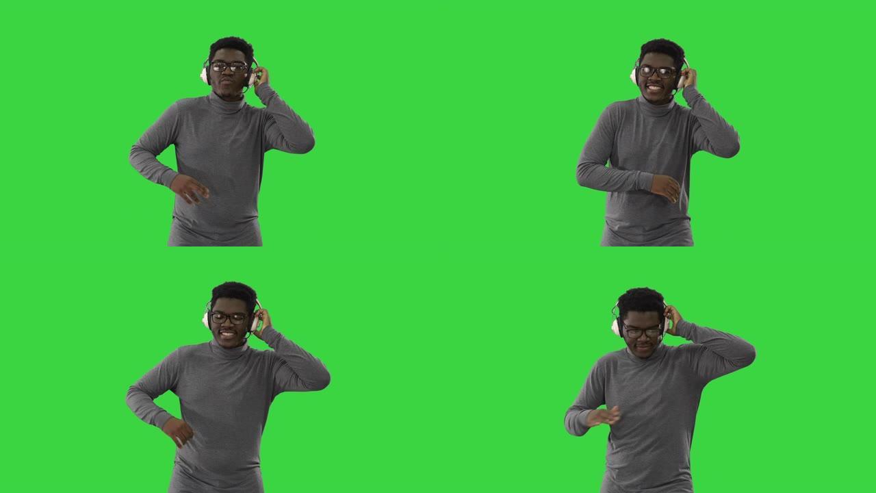 微笑的非洲裔美国男子用一只手拿着耳机，对着音乐开槽，在绿色屏幕上对着相机行走，色键