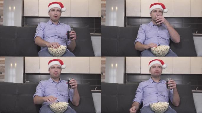 戴着圣诞帽的认真专心的家伙在房间里吃爆米花，喝sida和看电视的视频。用遥控器切换频道。圣诞节或新年