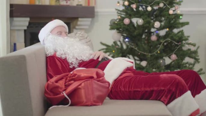 穿着圣诞老人服装的白人老人睡在圣诞树背景下的沙发上。疲惫的家伙在家艰难的一天后休息。
