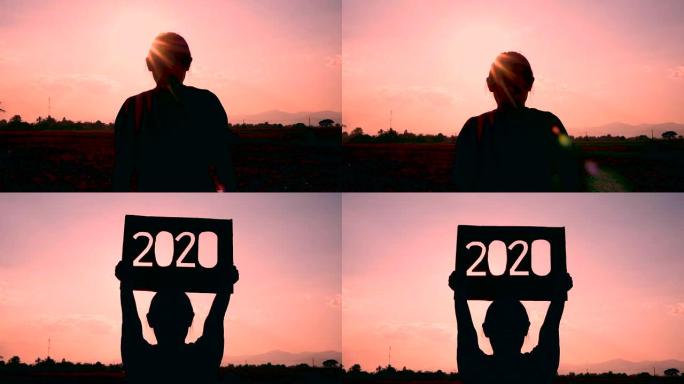 手拿着回收纸板的剪影与日落天空背景上的2020文本。新年和庆祝活动的概念。