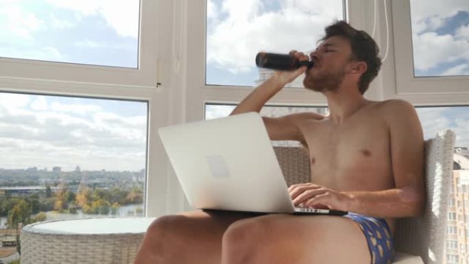 傲慢可怜的家伙穿着内裤坐在现代豪华的阳台上，玩电子游戏，喝啤酒。年轻的醉酒男性看着显示器，喝着酒