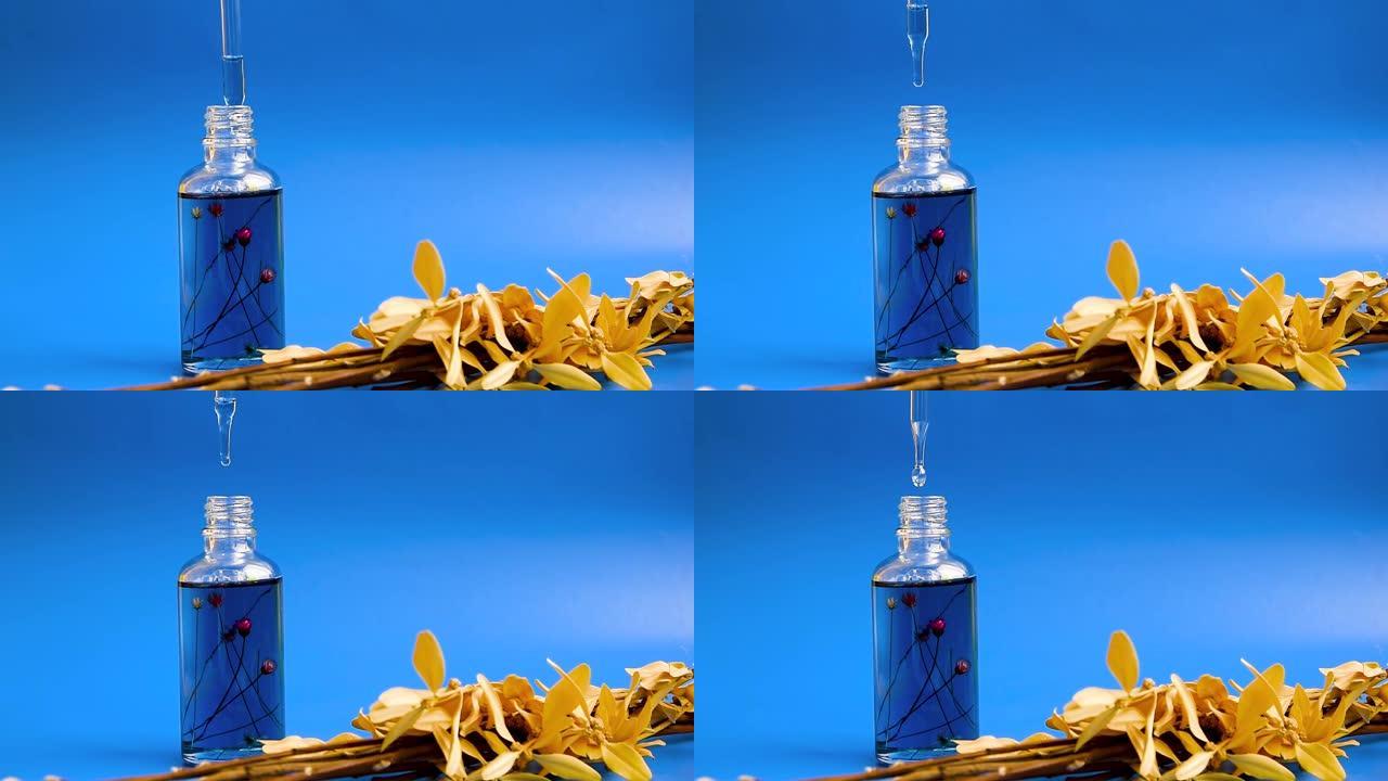 女性手中的滴管玻璃瓶模型，蓝色背景，干花。化妆品移液器上的油滴掉落，