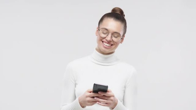 年轻快乐迷人的欧洲女孩穿着白色毛衣，手里拿着智能手机微笑着笑，孤立在工作室灰色背景上