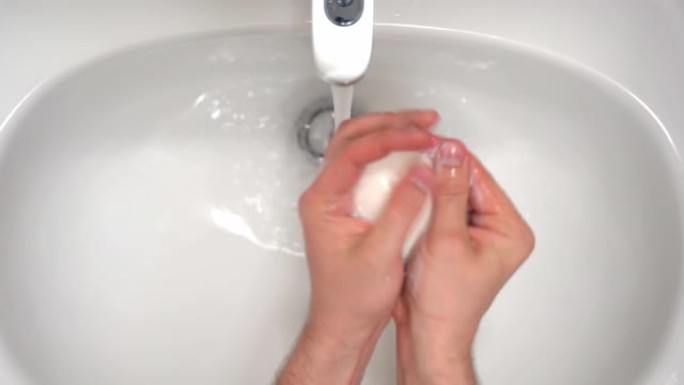 干净的白色浴室水龙头的俯视图，水龙头有流动的水，男人在温水下用肥皂清洁手，用于防止微生物膨胀的消毒肥