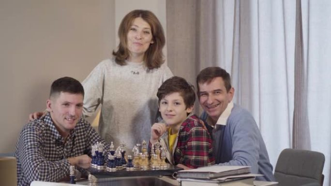 四个不同年龄的高加索人看着相机微笑。快乐的小男孩，他的父亲和祖父母晚上在室内下棋。快乐，休闲，生活方