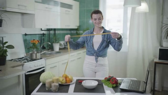 良好的饮食，年轻快乐的女人站在厨房旁边的桌子上健康的食物测量她的腰部用卷尺减肥成功后