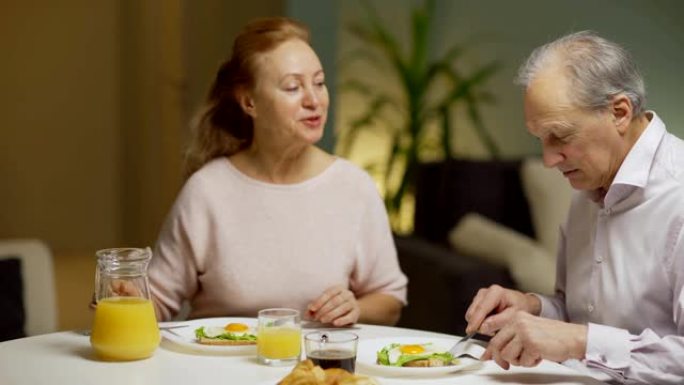 用一壶橙汁和丈夫一起吃鸡蛋作为早餐，向上倾斜妻子的中等镜头。高级夫妇坐在家里吃饭时兴高采烈地聊天
