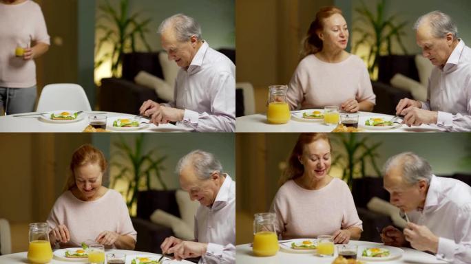 用一壶橙汁和丈夫一起吃鸡蛋作为早餐，向上倾斜妻子的中等镜头。高级夫妇坐在家里吃饭时兴高采烈地聊天