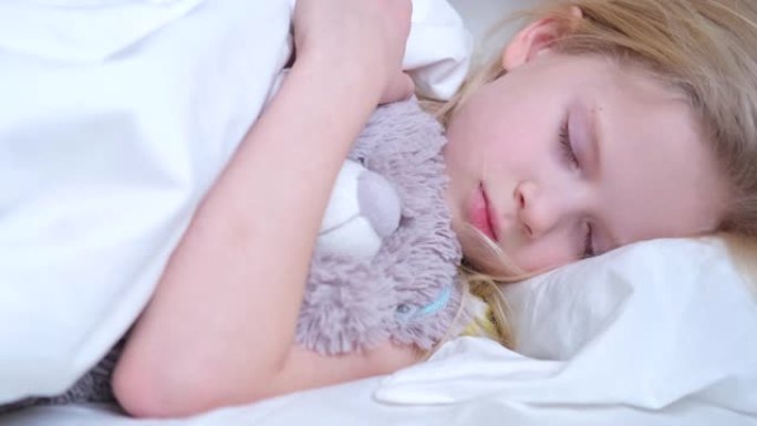 熟睡的小女孩在梦里拥抱一只泰迪熊。漂亮的金发孩子一大早就睡在床上，穿着白色床上用品。4k镜头