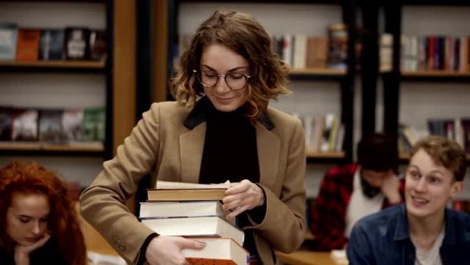 一个穿着棕色夹克和眼镜的女孩的慢动作镜头，她从书架上拿走了一堆书。其他学生坐着，在大学、大学图书馆学