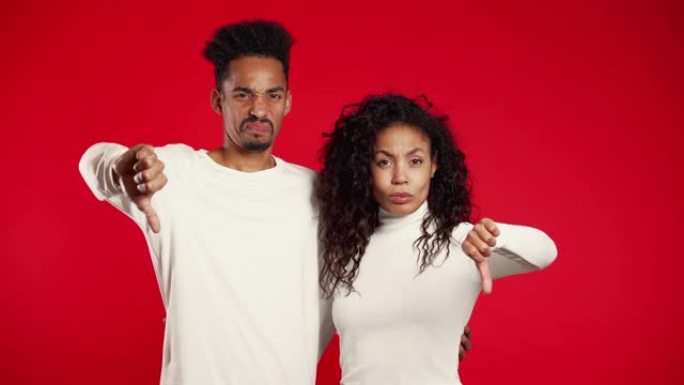 年轻的非洲夫妇站在红色的演播室背景上，表达不满，并在镜头前展示拇指向下的手势。带有不喜欢的迹象的男人