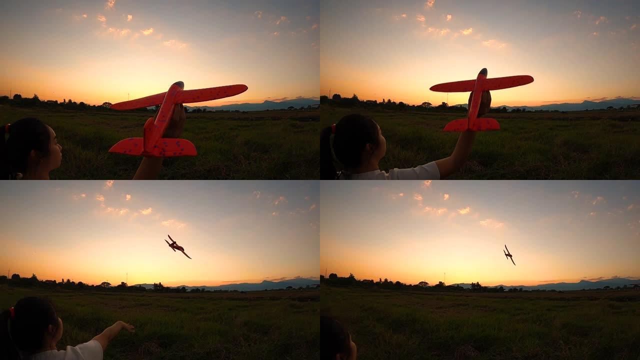 一个快乐的年轻女子的慢动作，手里拿着一架飞机，在田野上的日落背景下向前投掷。