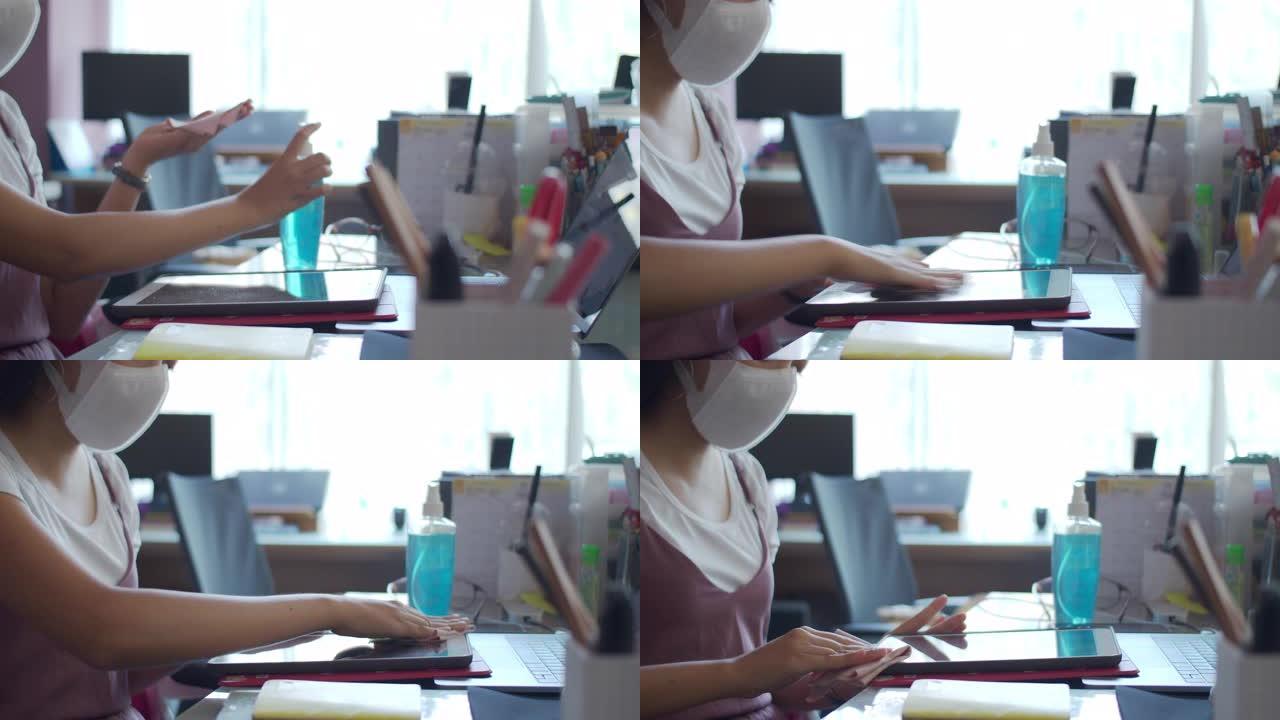 戴口罩的亚洲女商人在家庭办公室后台开始工作之前，使用酒精为基础的洗手液喷雾清洁平板电脑和智能手机。洗