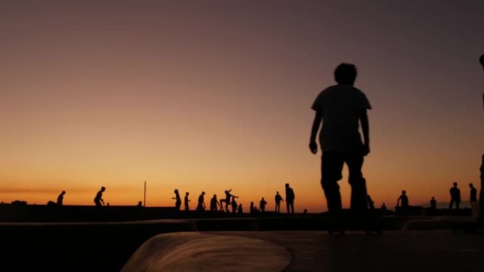 年轻跳跃滑板手骑长板的剪影，夏天的日落背景。加州洛杉矶威尼斯海洋海滩滑板公园。极限公园滑板坡道上的青