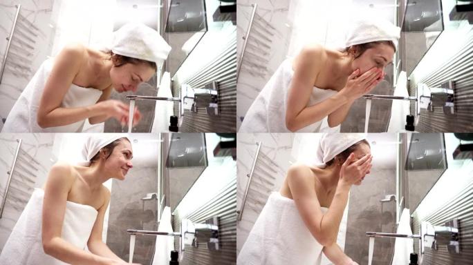 一名妇女站在水槽附近的浴室里，头上穿着白毛巾，用水龙头里的水洗脸。看着镜子的倒影和微笑。低角度视图