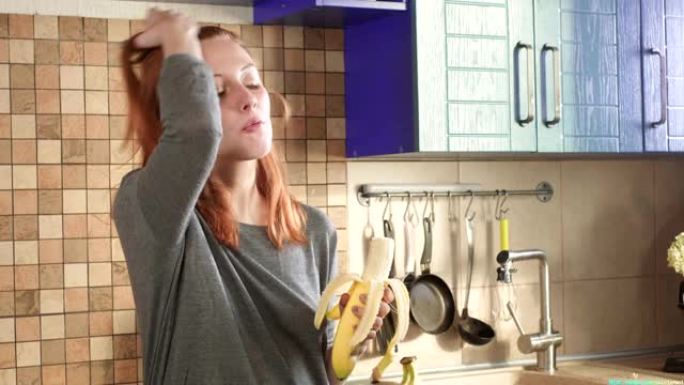 迷人的饥饿的红发女孩在周末的清晨在厨房里吃一个大香蕉。清洗果皮。健康饮食，正确的生活方式。运动早餐。