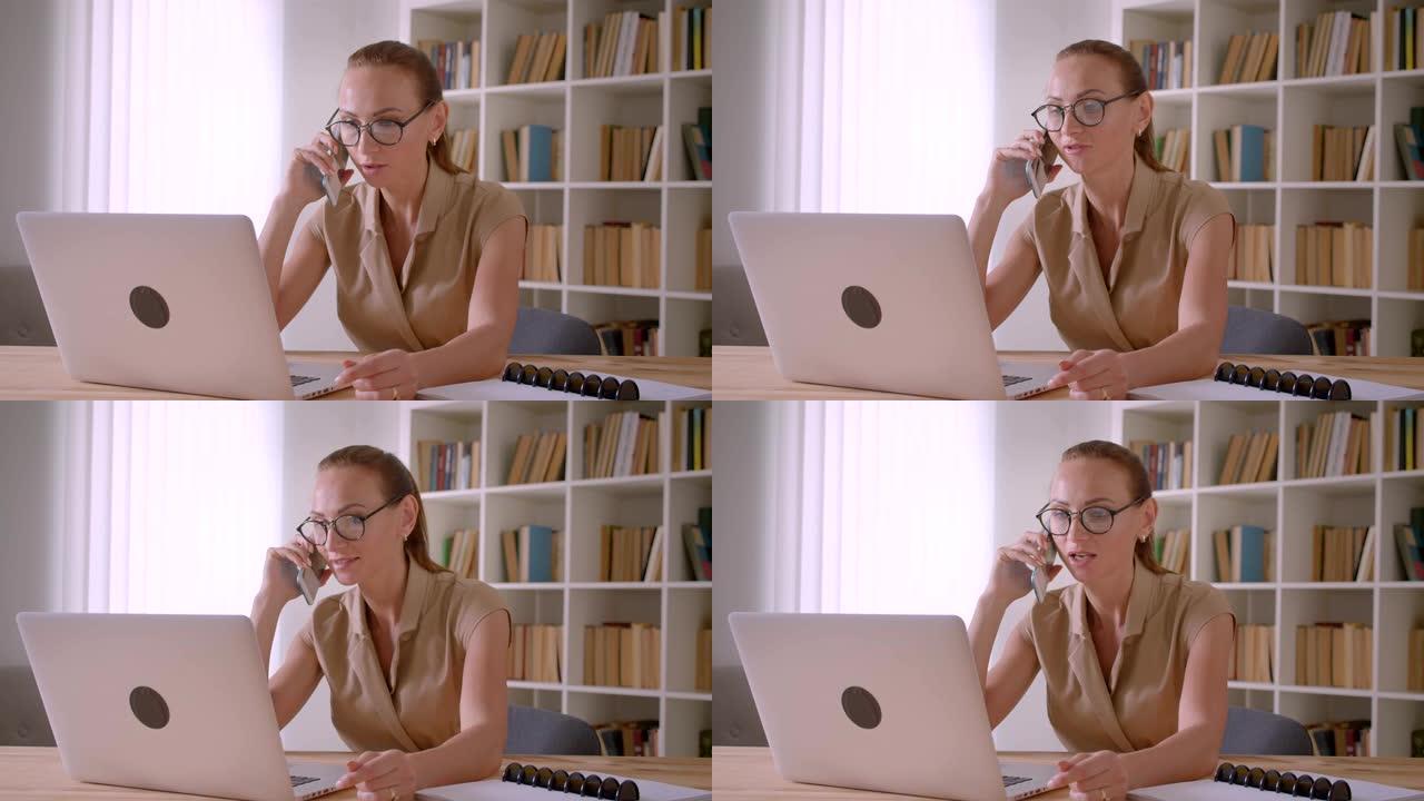 戴眼镜的白人女商人在手机上聊天，在办公室里把手表放在笔记本电脑上。