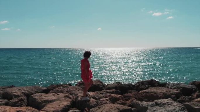 可爱的女士穿着红色连衣裙，随风飘动，在海上码头的岩石上行走，阳光反射在蓝色的海水上。塞浦路斯珊瑚海滩