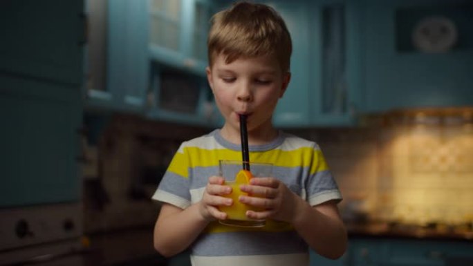 小男孩手里拿着一杯橙汁，用非塑料吸管慢动作喝。孩子在家里的厨房里享受新鲜的橙色饮料。