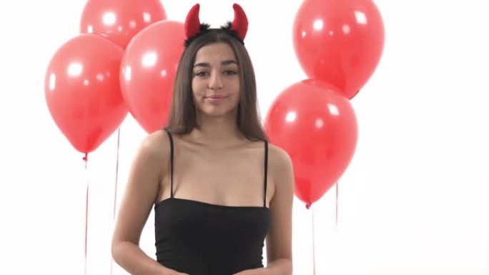一个戴着魔鬼头角和微笑的性感年轻女子的肖像。站在白色背景上的红色气球前的女人